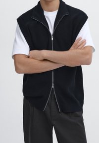 Mörkstickad väst med korta ärmar, hög krage och dragkedja fram. Buren över en vit t-shirt, matchad med mörka byxor. Enkel design.
