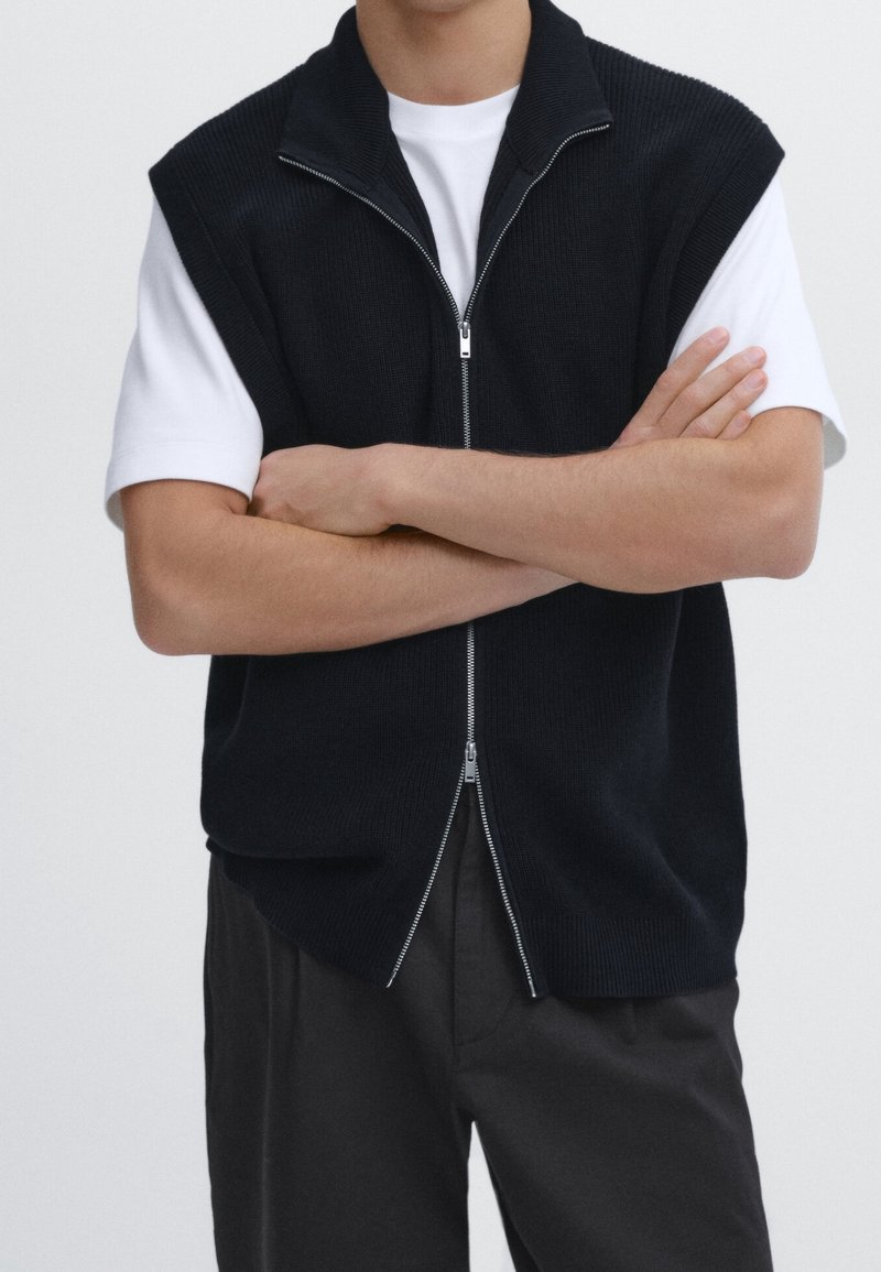 Mörkstickad väst med korta ärmar, hög krage och dragkedja fram. Buren över en vit t-shirt, matchad med mörka byxor. Enkel design.