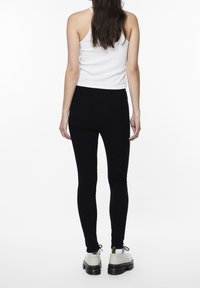 Kvinna som står vänd bort, iförd en vit linne, svarta skinny jeans, vita strumpor och beige plattformsskor med svarta sulor.