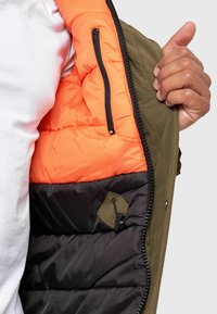 Chaqueta verde oliva con un forro interior acolchado naranja. Presenta bolsillos con cremallera negra y una capa exterior texturizada negra.