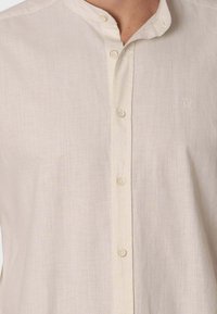 Camisa abotonada color beige claro, hecha de tela texturizada, con cuello alzado, botones visibles y un pequeño logotipo bordado en el pecho.