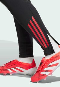 Zwarte sportbroeken met een rode drie-strepenaccent, voorzien van zijritsen. Felrode voetbalschoenen met witte strepen en een gestructureerde bovenkant.