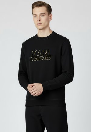 Młody mężczyzna ubrany w czarną bluzę Karl Lagerfeld z wypukłym złotym napisem, stojący na jednolitym jasnoszarym tle.