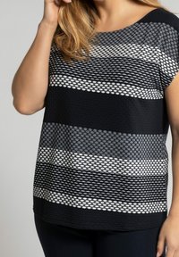 Femme portant un haut texturé à rayures noir, blanc et gris à manches courtes, avec une main touchant ses cheveux.