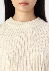 Maglione lavorato a maglia color crema con collo rotondo a coste. La trama è a maglia aperta, con sottili variazioni di luce e ombra.