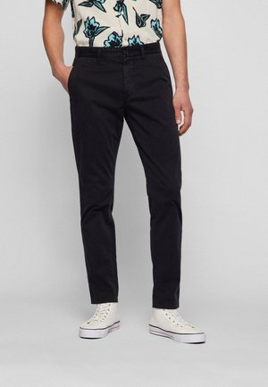 Homme portant un pantalon noir slim, des baskets blanches montantes et une chemise à manches courtes boutonnée avec un imprimé floral bleu.