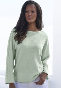 Hellgrünes, strukturiertes Langarmshirt mit rundem Ausschnitt und leichtem Saum. Verfügt über Knopfdetails an den Bündchen. Getragen mit weißen Hosen.