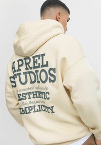 Cream-colored hoodie with a spacious hood, featuring dark gray text on the back. Text includes brand name and phrases in varying sizes.