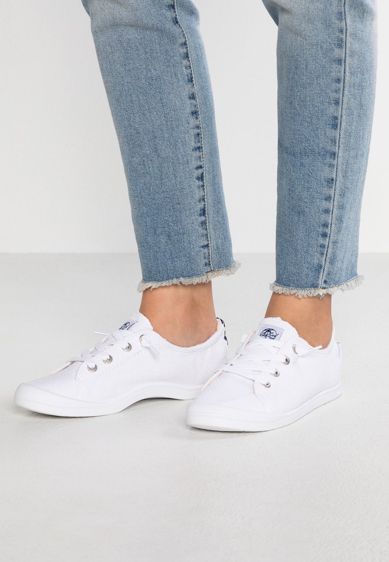 Jambes portant un jean bleu clair avec des ourlets effilochés et des baskets en toile blanches à lacets sur un sol clair uni.
