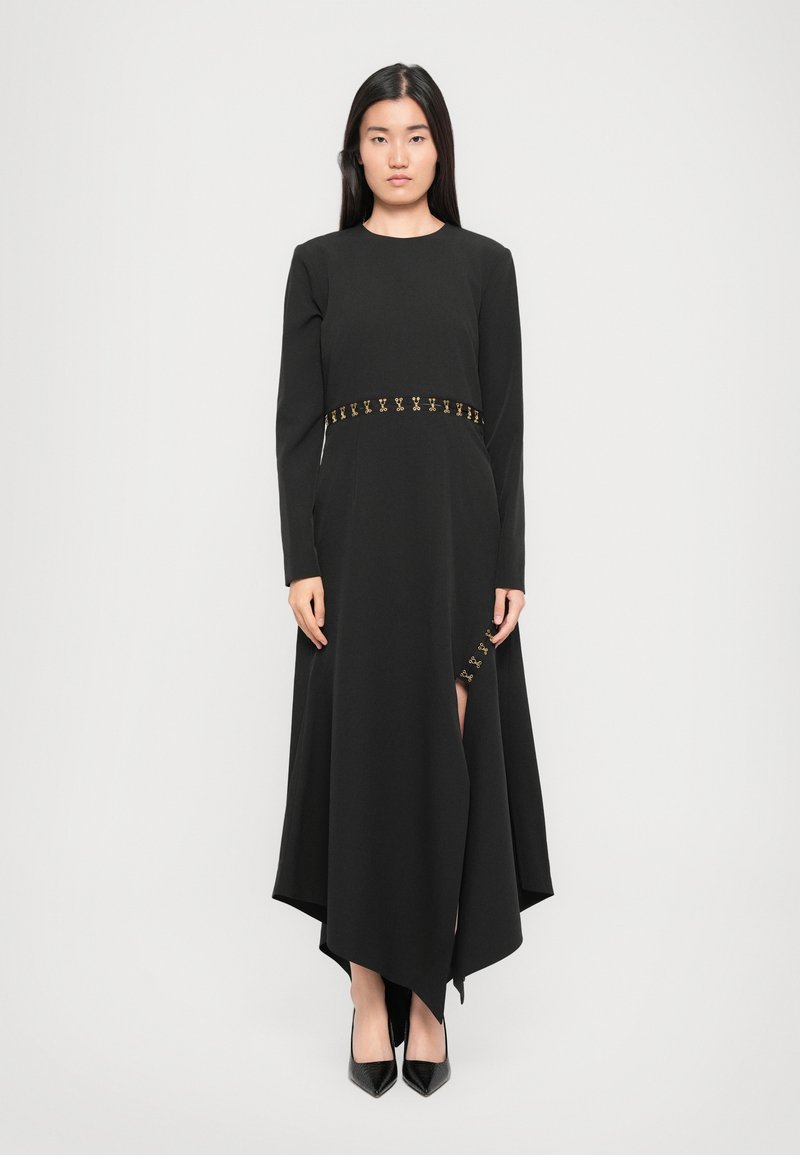 Black long-sleeve dress with asymmetric hem, featuring gold accents at the waist and side slit. Smooth fabric texture.