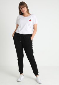 Balta T-krekls ar sarkano ķiršu grafiku, kombinēts ar melniem sporta biksēm ar baltām sānu svītrām, abi izgatavoti no mīkstas kokvilnas auduma.