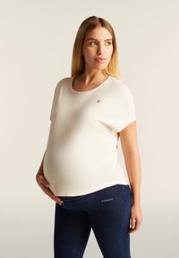 Top premaman color crema a maniche corte con scollatura rotonda, abbinato a leggings neri elasticizzati con un logo in vita.