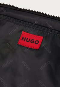 Melna auduma maisiņš ar sarkanu taisnstūrveida uzlīmi, kurā rakstīts "HUGO" melnā krāsā. Virsma ir gluda, un tam ir rāvējslēdzēja aizdare.