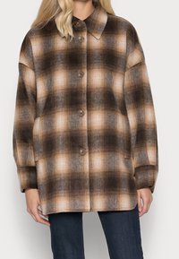 Ruutukuvioinen ruskea ja vaaleanruskea takki, joka on valmistettu teksturoidusta kankaasta, sisältää napituskiinnityksen, kauluksen ja pitkät hihat.