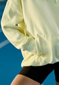 Gele katoenen hoodie met een losse pasvorm en een zak aan de voorkant, gecombineerd met zwarte fietsshorts, op een blauwe achtergrond.