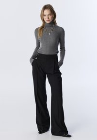 Grauer Rollkragenpullover mit subtilen Glitzereffekten, kombiniert mit hochgeschnittenen schwarzen Hosen, die über Schlaufen verfügen, und eleganten schwarzen Schuhen.