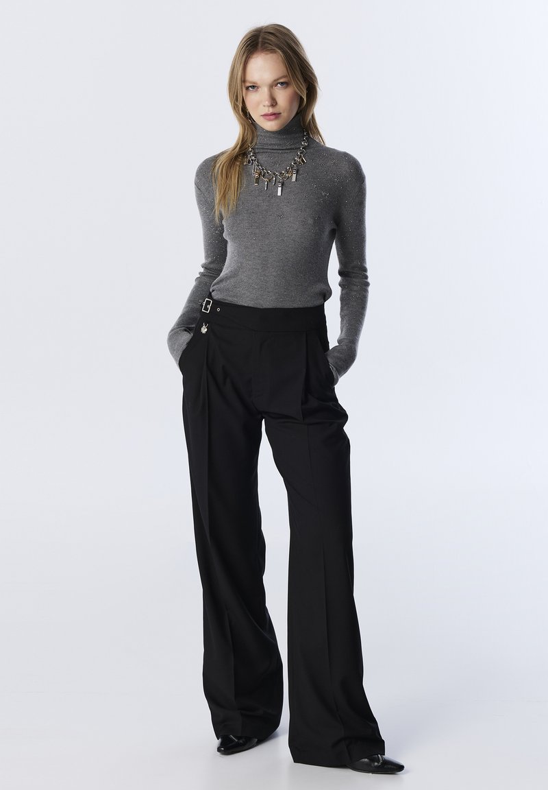 Grauer Rollkragenpullover mit subtilen Glitzereffekten, kombiniert mit hochgeschnittenen schwarzen Hosen, die über Schlaufen verfügen, und eleganten schwarzen Schuhen.