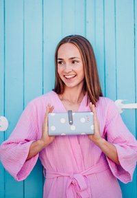 Light blue wallet with large white polka dots, featuring a gray button closure and a small logo. Held in front of a turquoise wooden background.