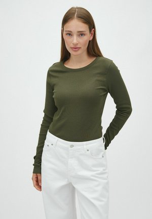 Junge Frau mit glattem hellbraunem Haar trägt ein olivgrünes Langarmtop und hoch taillierte weiße Hosen vor einem einfachen Hintergrund.