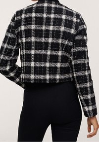 Sort-hvid ternet cropped jakke med et tekstureret stof, der har en krave og lange ærmer, vist fra ryggen.