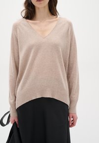 Helles beigefarbenes Strickpullover mit V-Ausschnitt, geripptem Saum und überschnittenen Schultern, kombiniert mit einem schwarzen Rock. Weiche Textur, die im Material sichtbar ist.