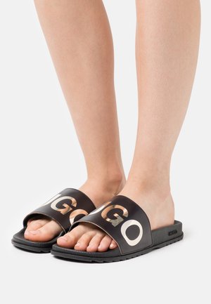Schwarze Sandalen mit breiten Riemen und einem auffälligen weißen Logo. Die strukturierte Gummisohle sorgt für Grip, während das Obermaterial glatt und kontrastreich ist.