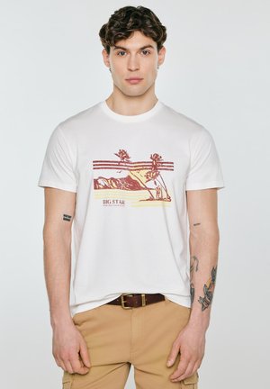 Młody mężczyzna w białej koszulce z nadrukiem gór i palm, beżowych spodniach, brązowym pasku i widocznymi tatuażami na ramionach, stoi nieruchomo.