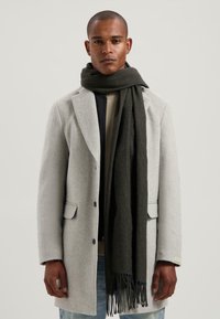 Man in een lichtgrijze wollen jas, met een donkergrijze sjaal om zijn nek, een Beige shirt en lichtblauwe jeans, staand tegen een neutrale achtergrond.