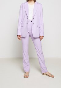 Fialový přiléhavý blazer se dvěma předními kapsami a černými knoflíky, doplněný o úzké kalhoty ve stejném odstínu. Nosený s lehkým topem a sandály.