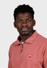 Pull-over en polaire rose clair à fermeture éclair quart, avec un petit écusson logo représentant un éléphant. Texture douce avec un col et des détails de fermeture éclair.