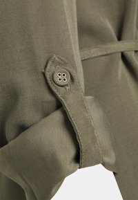 Tessuto verde oliva con una texture liscia. Dettagli notevoli includono una manica arrotolata con un'etichetta cucita e una chiusura a bottone metallico.