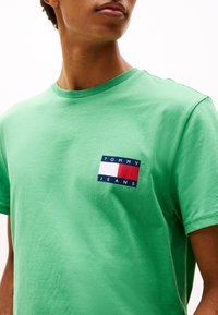 Green cotton t-shirt with a crew neck, featuring a small, rectangular Tommy Jeans logo patch in navy, white, and red on the left chest.