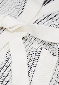 Närbild av vitt stickat tyg med svarta detaljer och ett tjockt vitt stickat bälte som är knutet löst över det.