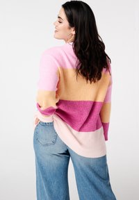 Pull en tricot avec des rayures horizontales roses, pêches et lavande. Présente une coupe ample et une texture côtelée, porté avec un jean taille haute.