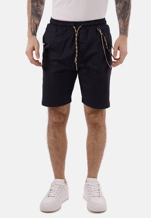Pantaloni corti blu navy con cintura elastica, cordino regolabile, tasche laterali e un leggero lucido; abbinati a sneakers bianche.
