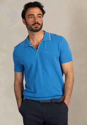 Man met baard, gekleed in een felblauw, kort mouws overhemd met kraag en donkere broek, staand met handen in de zakken tegen een effen achtergrond.