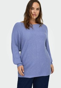 Hele sinine soonikuga kampsun lõdva lõikega, lai paadi kaelus ja pikad varrukad, kantud tumedate pükste kohal, millel on tekstuuriga viimistlus.