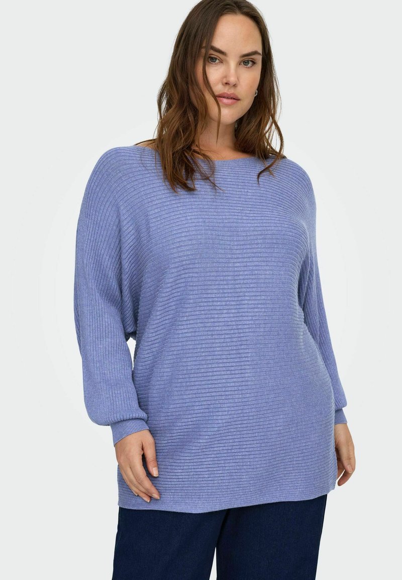 Hele sinine soonikuga kampsun lõdva lõikega, lai paadi kaelus ja pikad varrukad, kantud tumedate pükste kohal, millel on tekstuuriga viimistlus.