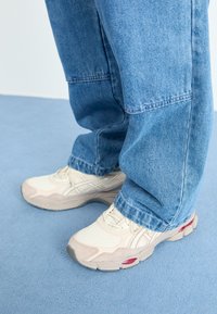 Beige träningsskor med texturerad mesh och mockadetaljer, med en gummisula som har en röd detalj. Bärs med ljusblå denimjeans.