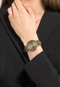 Ronde horloge met een matte olijfgroene band en een goudkleurige kast. De wijzerplaat heeft verhoogde uurmarkeringen en een zonnestraalpatroon.