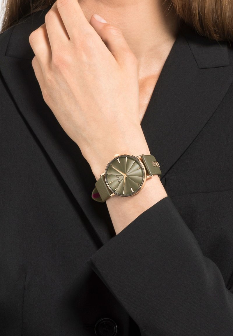 Ronde horloge met een matte olijfgroene band en een goudkleurige kast. De wijzerplaat heeft verhoogde uurmarkeringen en een zonnestraalpatroon.