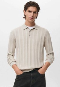 Camisa polo de manga larga a rayas beige con una tapeta de tres botones y puños de canalé, combinada con jeans oscuros, de pie frente a un fondo neutro.