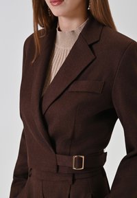 Bruine op maat gemaakte blazer met een subtiele textuur en een enkele borstzak, voorzien van een ceintuurdetail met een gouden gesp. Draagt over een geribbelde beige top.