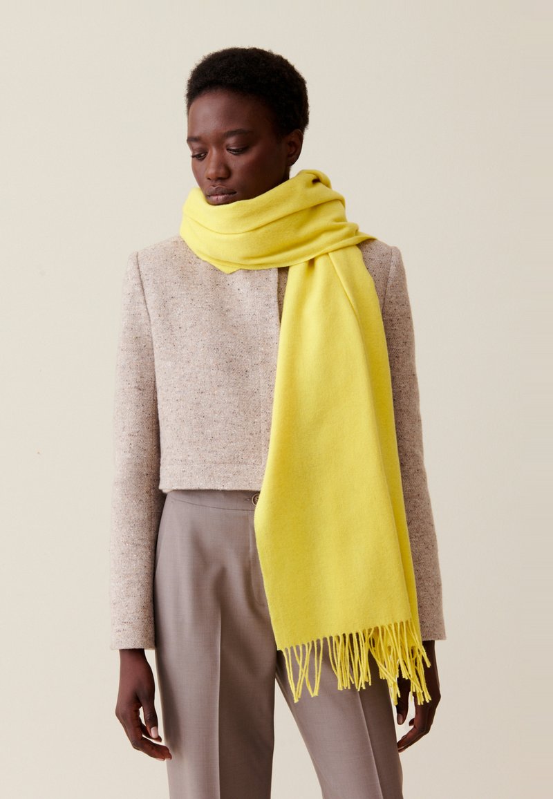 Gelber Schal aus weicher Wolle mit fransigen Kanten, um den Hals drapiert, kombiniert mit einer kurzen, gesprenkelten beigen Jacke.