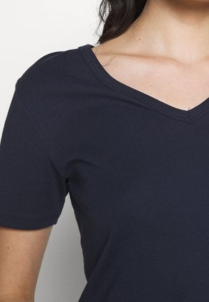 Kvinde iført en mørkeblå T-shirt med korte ærmer og V-hals, der viser skuldre og øvre bryst mod en ensartet lys baggrund.