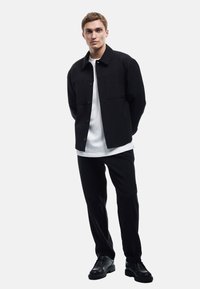 Veste noire à boutons sur un t-shirt blanc, accompagnée de pantalons noirs et de chaussures noires. Tissu lisse, design minimaliste et coupe classique.