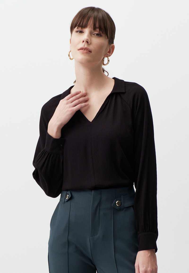 Blouse noire à manches longues avec un décolleté en V et un tissu texturé ; portée avec un pantalon vert taille haute orné de boutons décoratifs.
