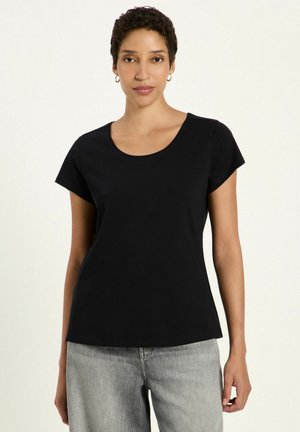 Woman with short curly hair wears a black short-sleeve top and light gray jeans, standing against a plain light background.