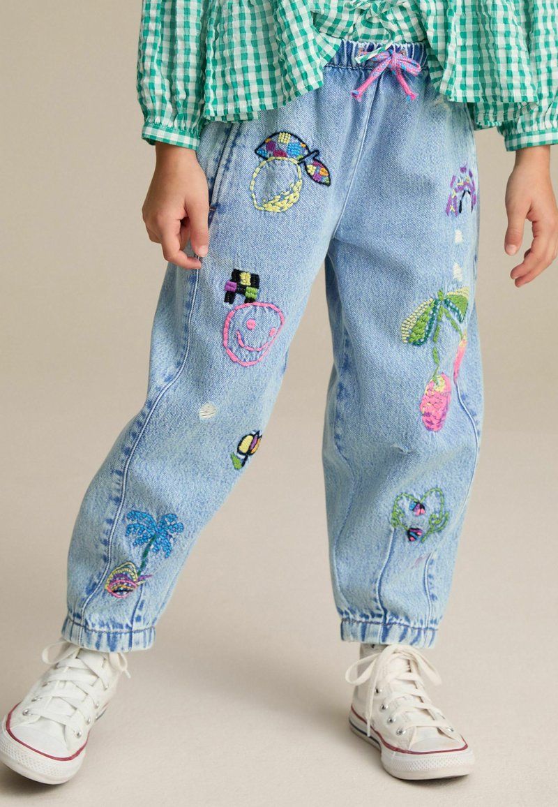 Barn iført lyseblå jeans med farverige broderede designs og en grønternede skjorte, står på en neutral baggrund.