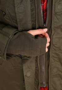 Ruka v hnědé palčáku s prsty částečně zapnutá do vrstvených olivově zelených a hnědých bund, s viditelnou červenou kostkovanou podšívkou.